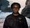 Artist John Akomfrah stands and looks directly towards the camera. He is smiling and wearing a dark denim jacket. He is standing in front of one of his films so the film is projected across him and the background behind him. The film shows a large rock in the sea with waves crashing on the sand.