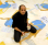 Artist N.S. Harsha sitting on his artwork whilst looking at the camera holding a paintbrush and cup of paint