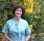 Chelsea Pettit is standing outside in front of a hedgerow. She is looking and smiling directly towards the camera. She has short brown curly hair and is wearing a blue and white dress. Out of focus, you can see there is a path with a black cat sitting on it.