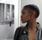 A woman called Cynthia MaiWa Sitei looks at a black and white photograph to her right-hand side. She has dark braided afro hair and is wearing a black leather jacket and long dangly earrings.