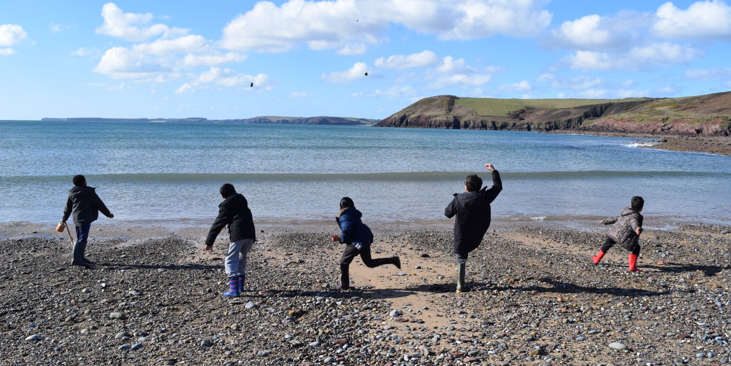 Five boys wearing wellies and coats are facing the sea and throwing rocks into the water. The rocks are mid-air. The sky is blue with light cloud and the coastline is visible into the distance.