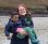A woman with orange hair and who is wearing glasses is holding a young boy in her arms. They are both smiling. They are stood on a beach and in the distance there is a grassy embankment.