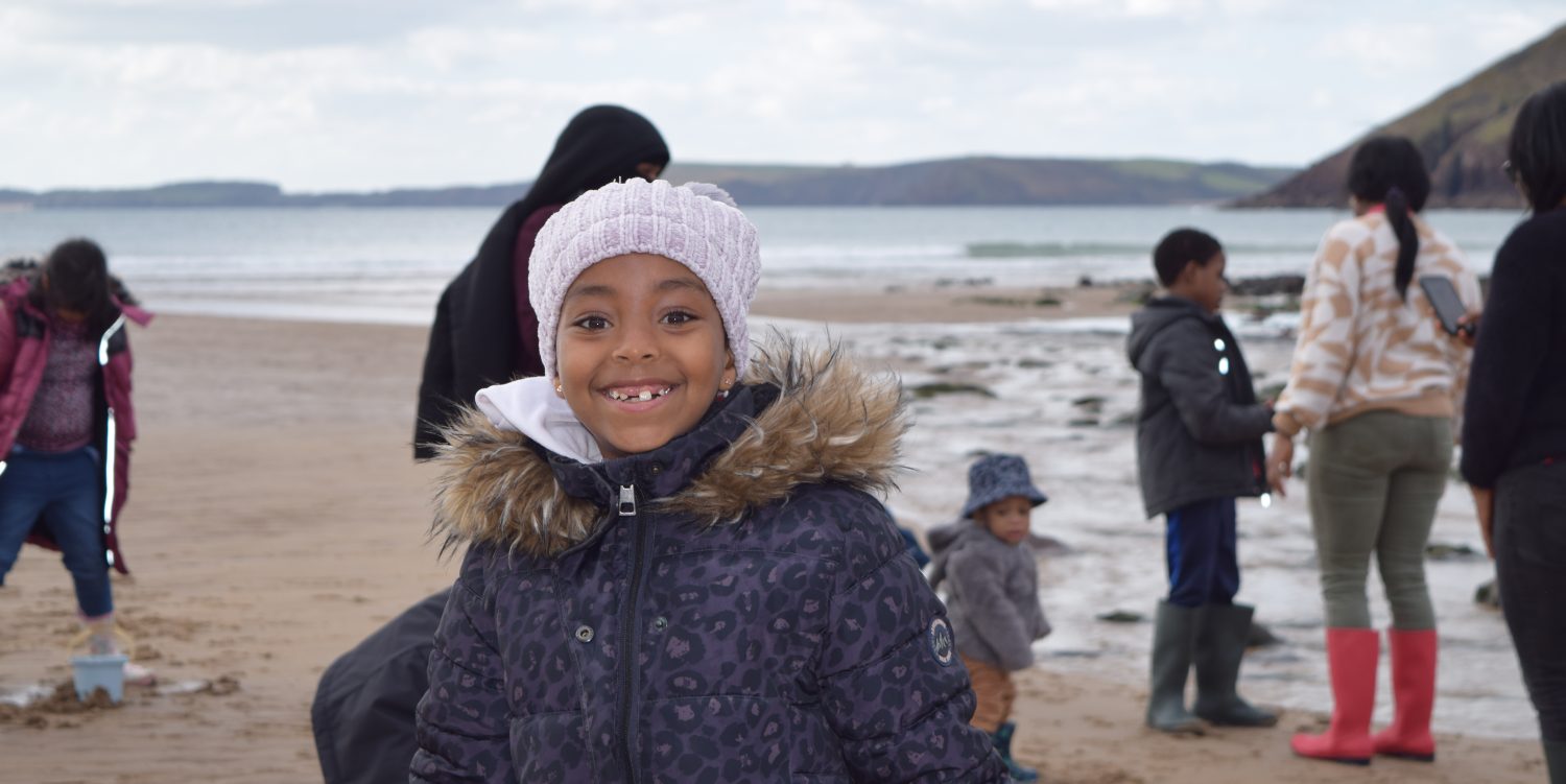 A young girl wearing a hat and a coat is smiling directly at the camera. Behind her other people are walking around on the beach.