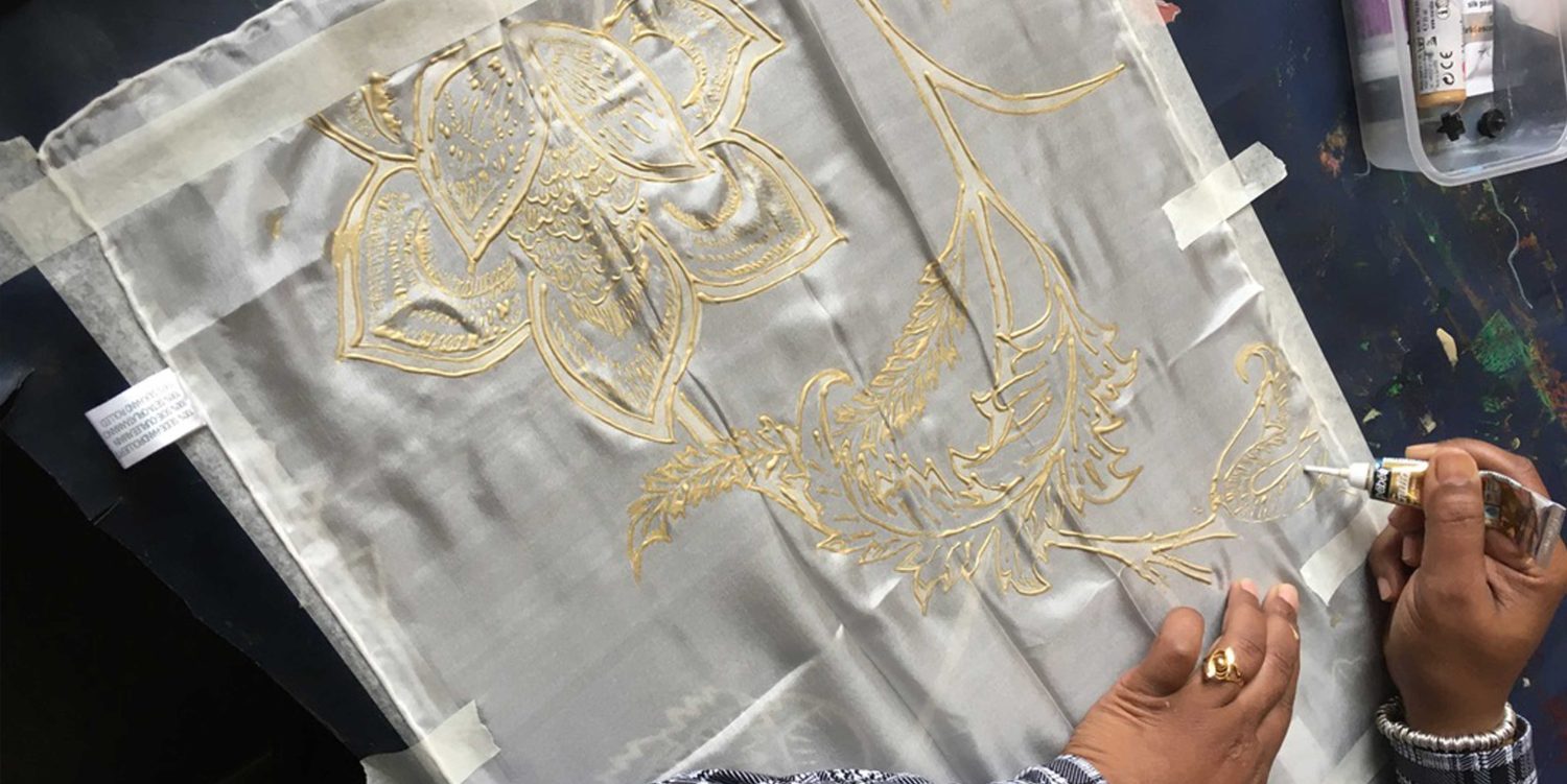 A persons hand is squeezing gold paint from a tube directly on to white fabric, creating a pretty, golden floral design.