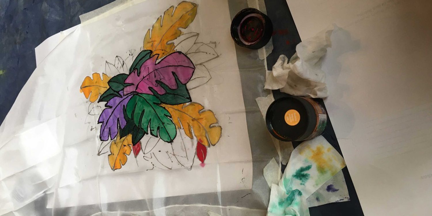 A colourful leaf design is painted on to white fabric.