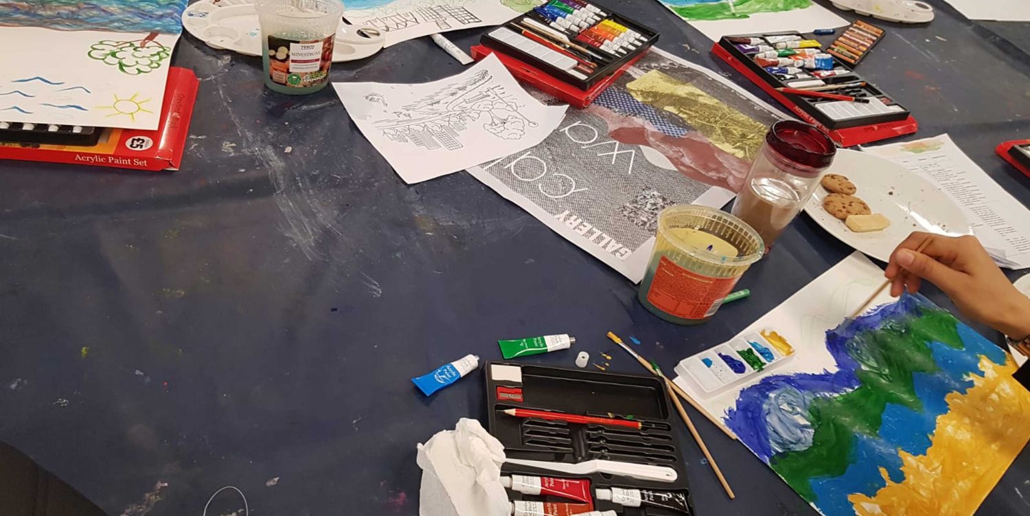 Around the edge of a table you can see that people are sat painting on white pieces of A4 paper. On the table are plenty of watercolour paints and palettes.