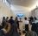 A group of people sat on chairs in a bright white room listening to a man wearing black talking and gesturing to the presentation on screen.
