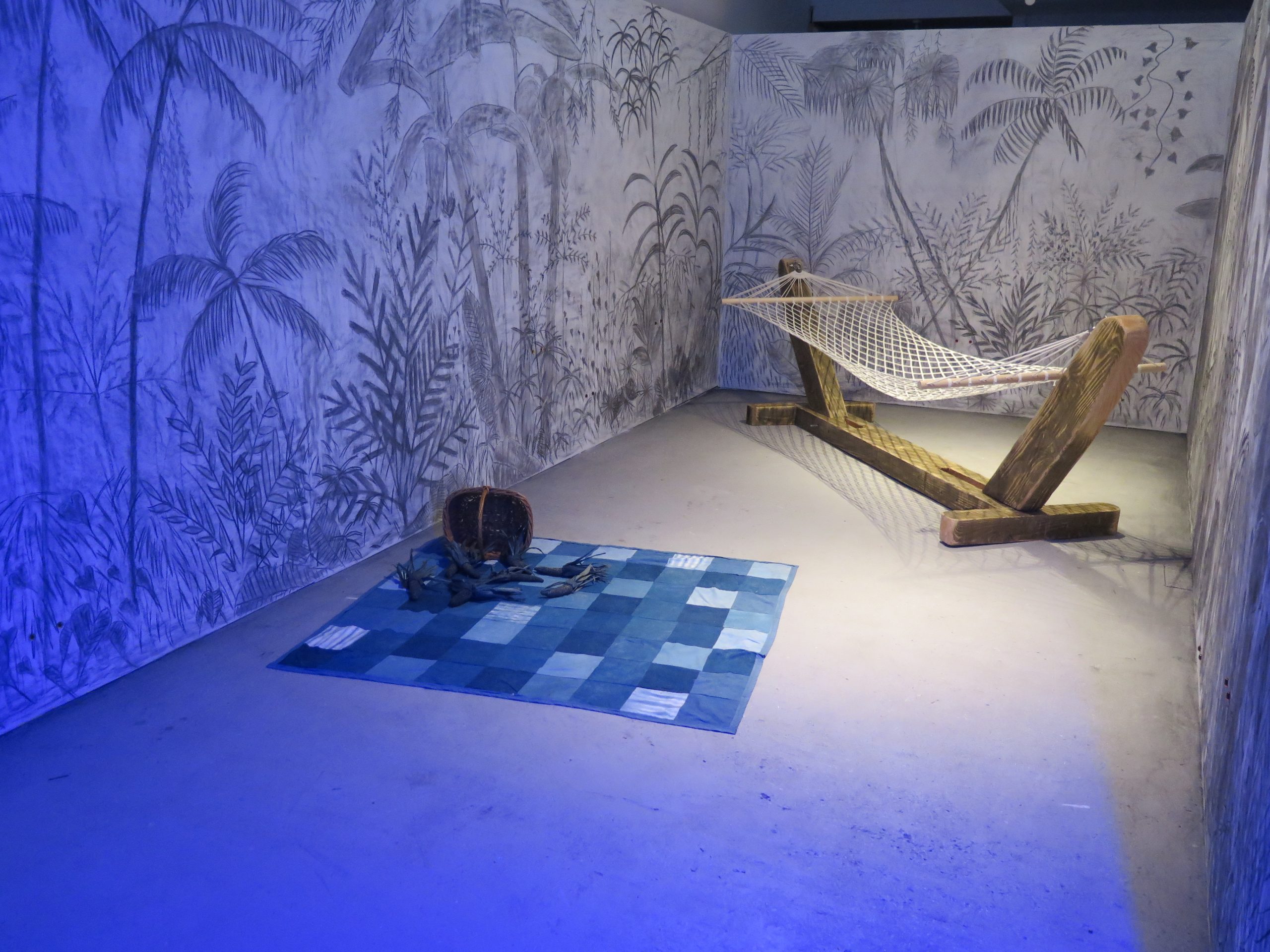 Daniel Trivedy – Between a Dream and a Nightmare. Charcoal drawings of trees and plants cover three white walls. Inside there is a hammock and on the floor there is a patchwork blanket which has been dyed indigo blue. On top of the blanket is a tipped-over basket with dyed blue corn falling out of it.
