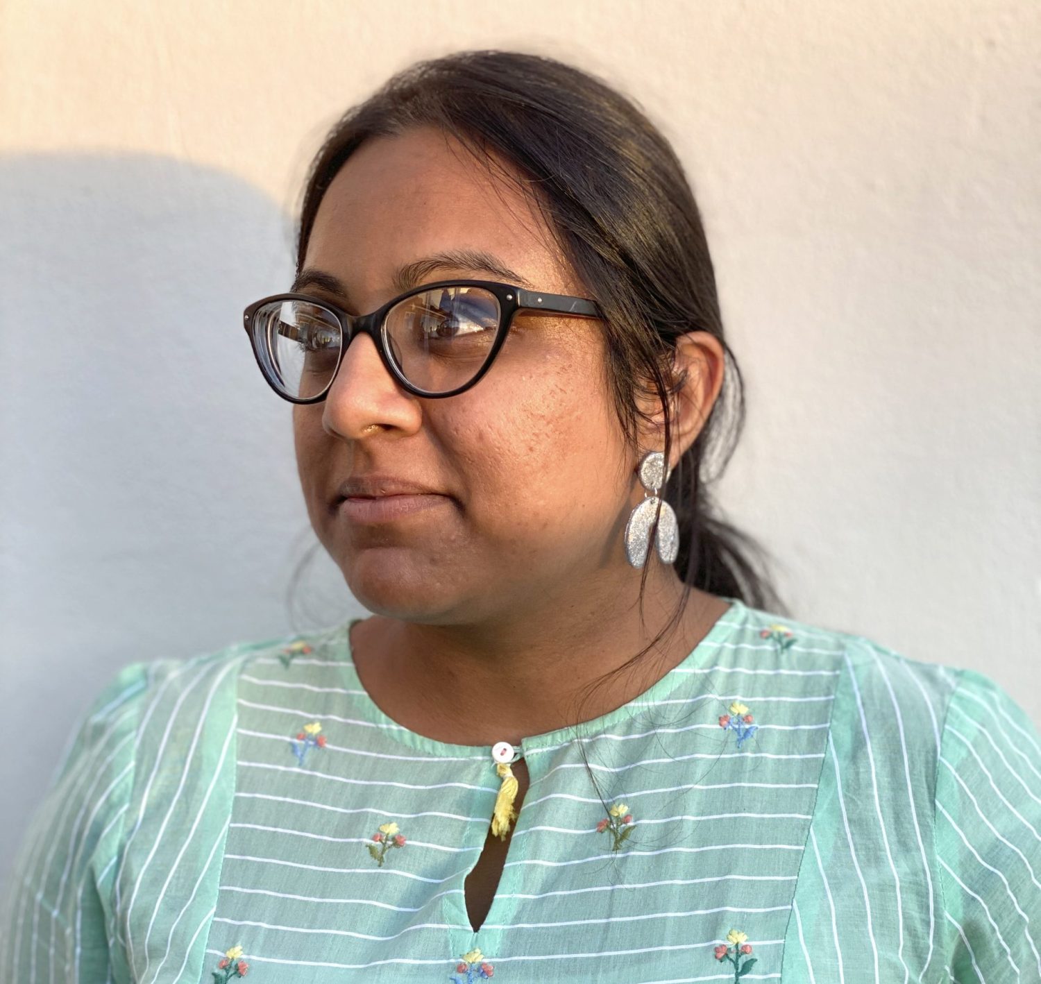 A close up photo of a woman wearing glasses and a pale green blouse with a floral print. Her face is slightly turned and she is looking in the same direction. She is in front of a plain white wall.