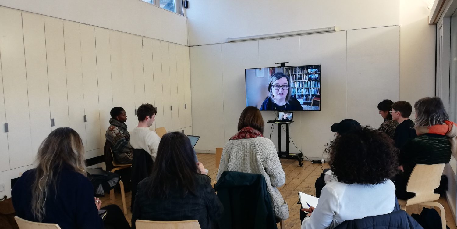A group of people sat on chairs in a bright white room listening to a woman wearing glasses who is talking them through zoom which is displayed on a screen in front of them.