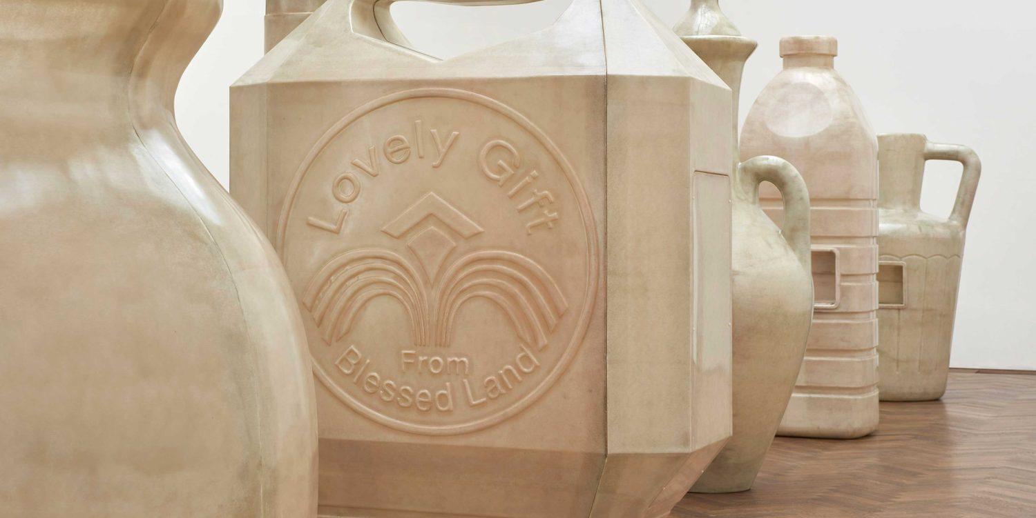 Five large fibreglass water vessel sculptures stood in a row in a large bright exhibition space. On the side of one of the sculptures it reads “Lovely Gift From Blessed Land”.