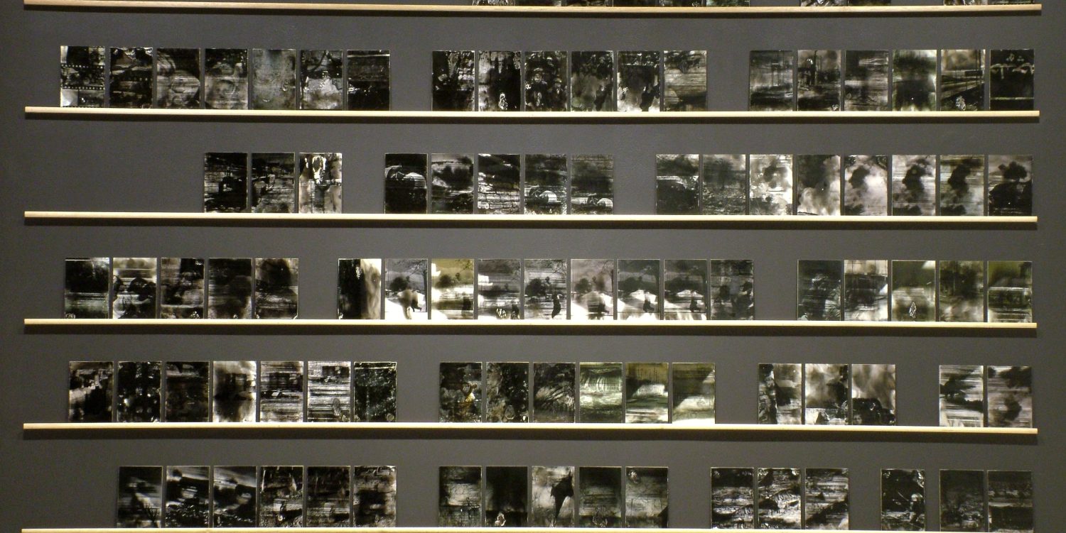 Installation view of multiple photos print treated with smoke (soot) mounted on six wooden shelves on a painted grey wall.