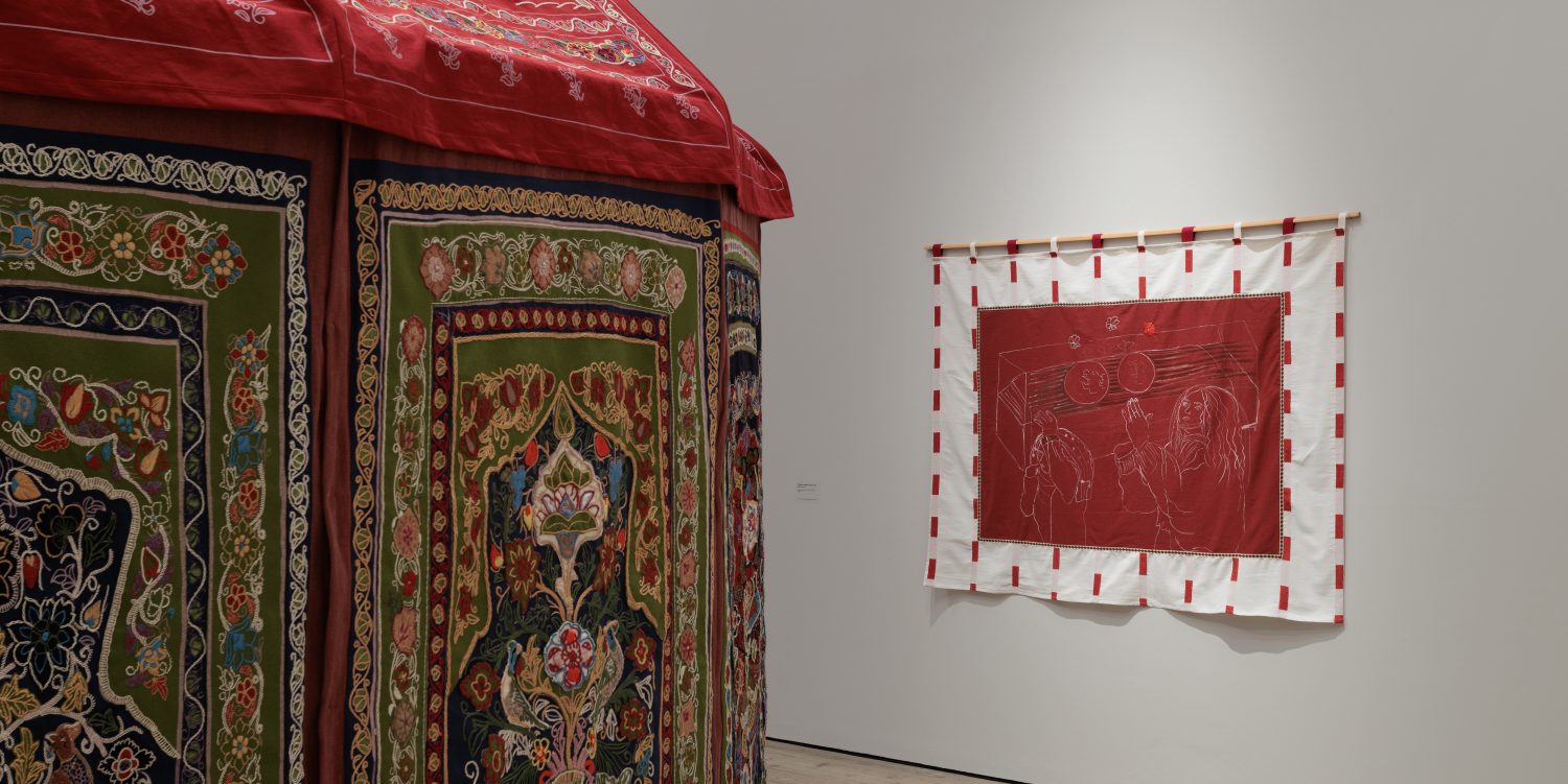 In the foreground you can see the corner of a circular red tent which is elaborately embroidered. In the background there is a red cotton and white wool artwork with ink and embroidery hung on a white exhibition wall.