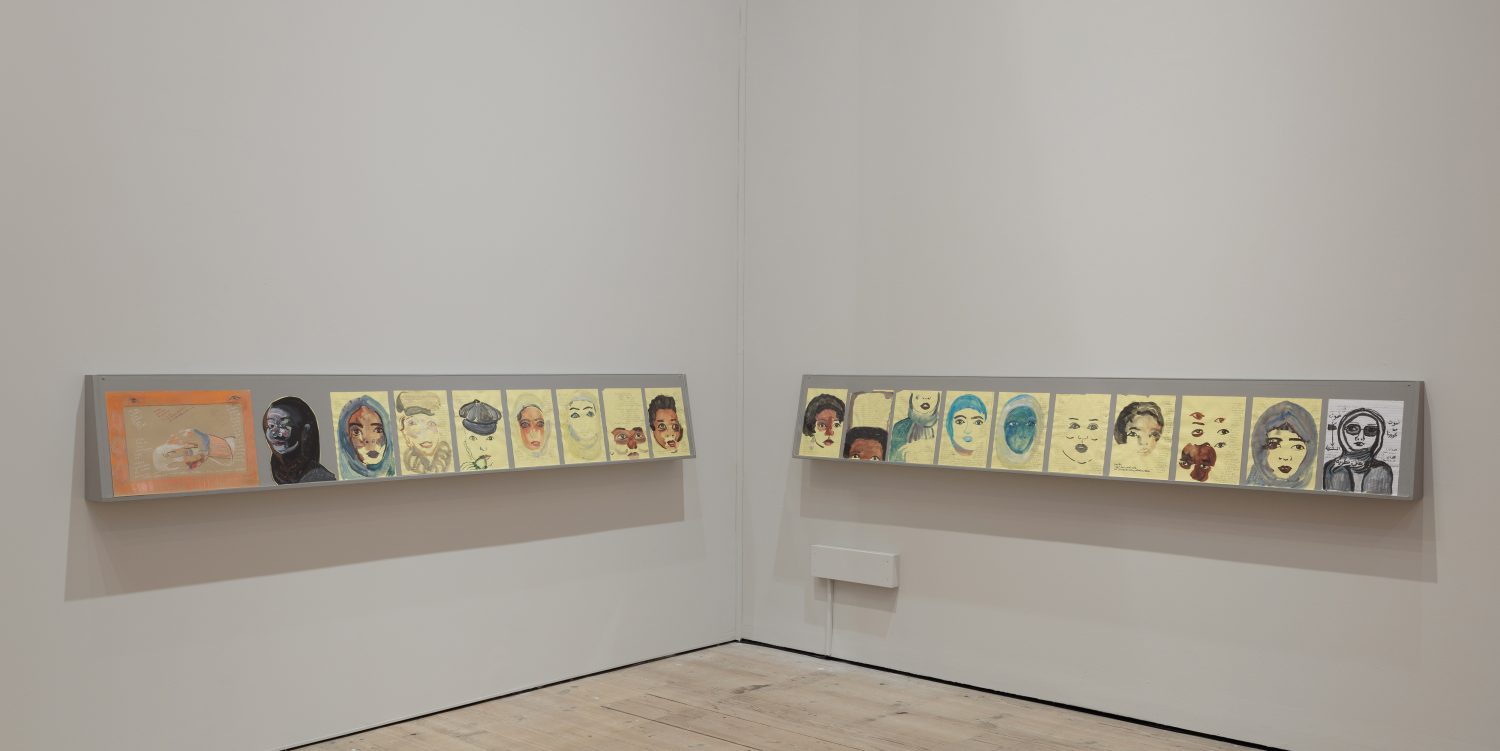 Multiple mixed media drawings on legal paper, mostly of women, are on display on long shelves in a white exhibition space.