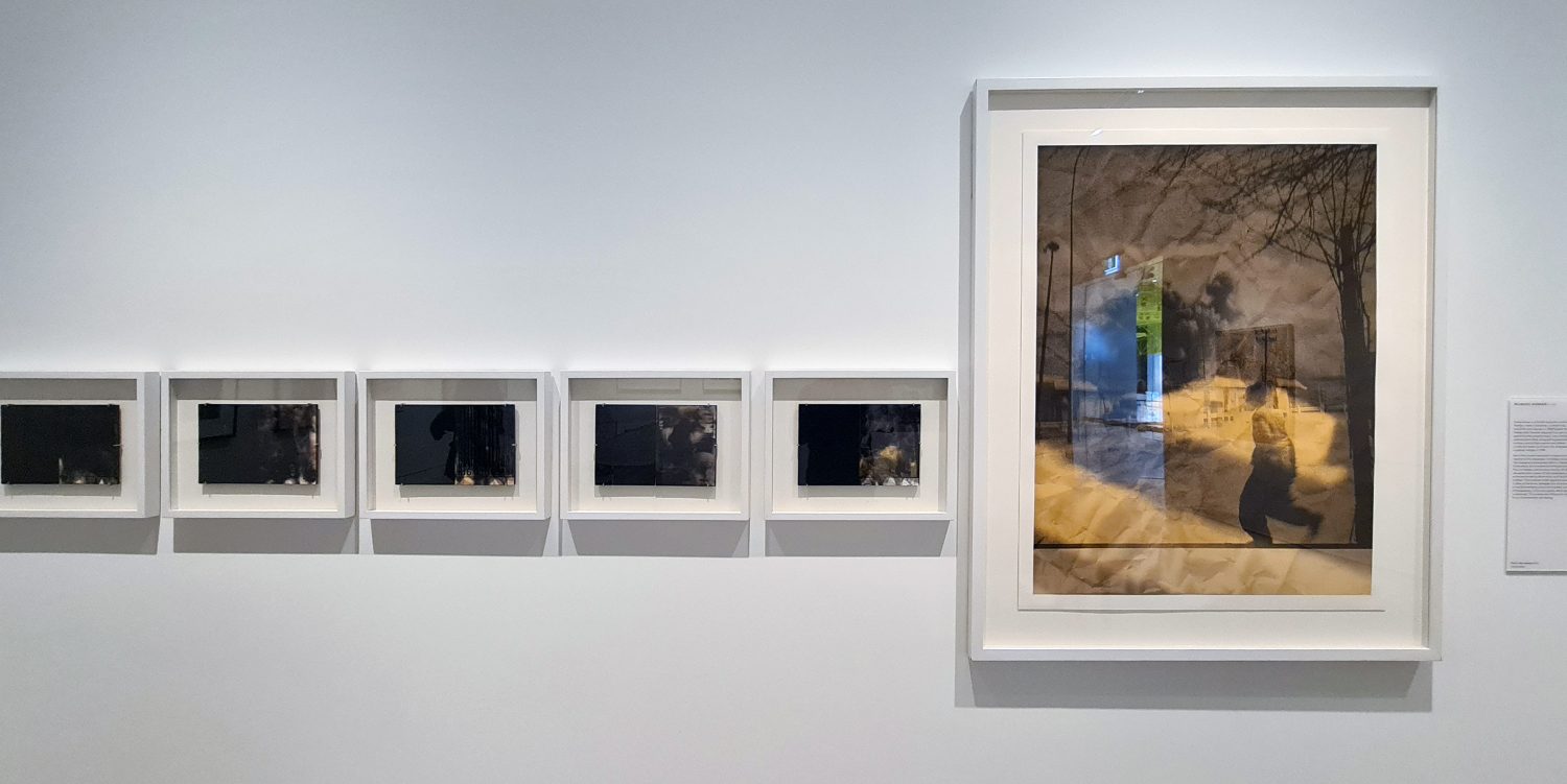 A row of five small, framed photographs treated with smoke. To the left is a larger print in a frame. They are secured to a white wall.