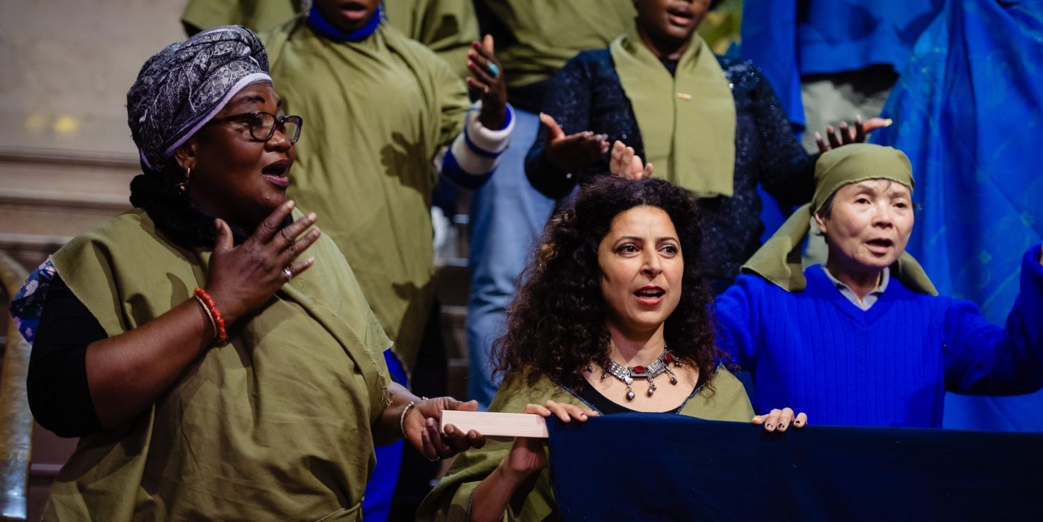 People in a choir wearing khaki and blue clothes are mid-song, using their hands to gesture.