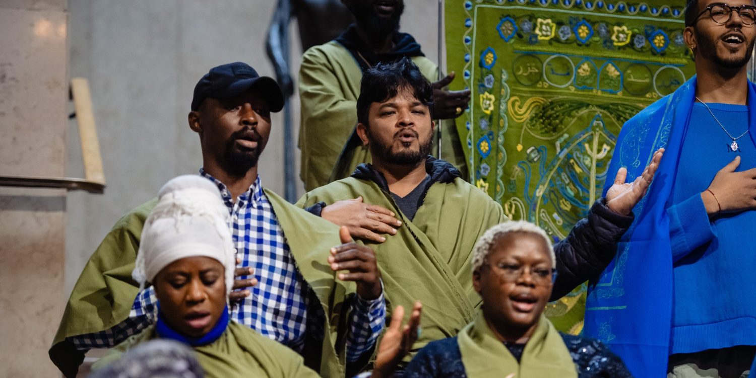 People in a choir wearing khaki and blue clothes are mid-song, using their hands to gesture.