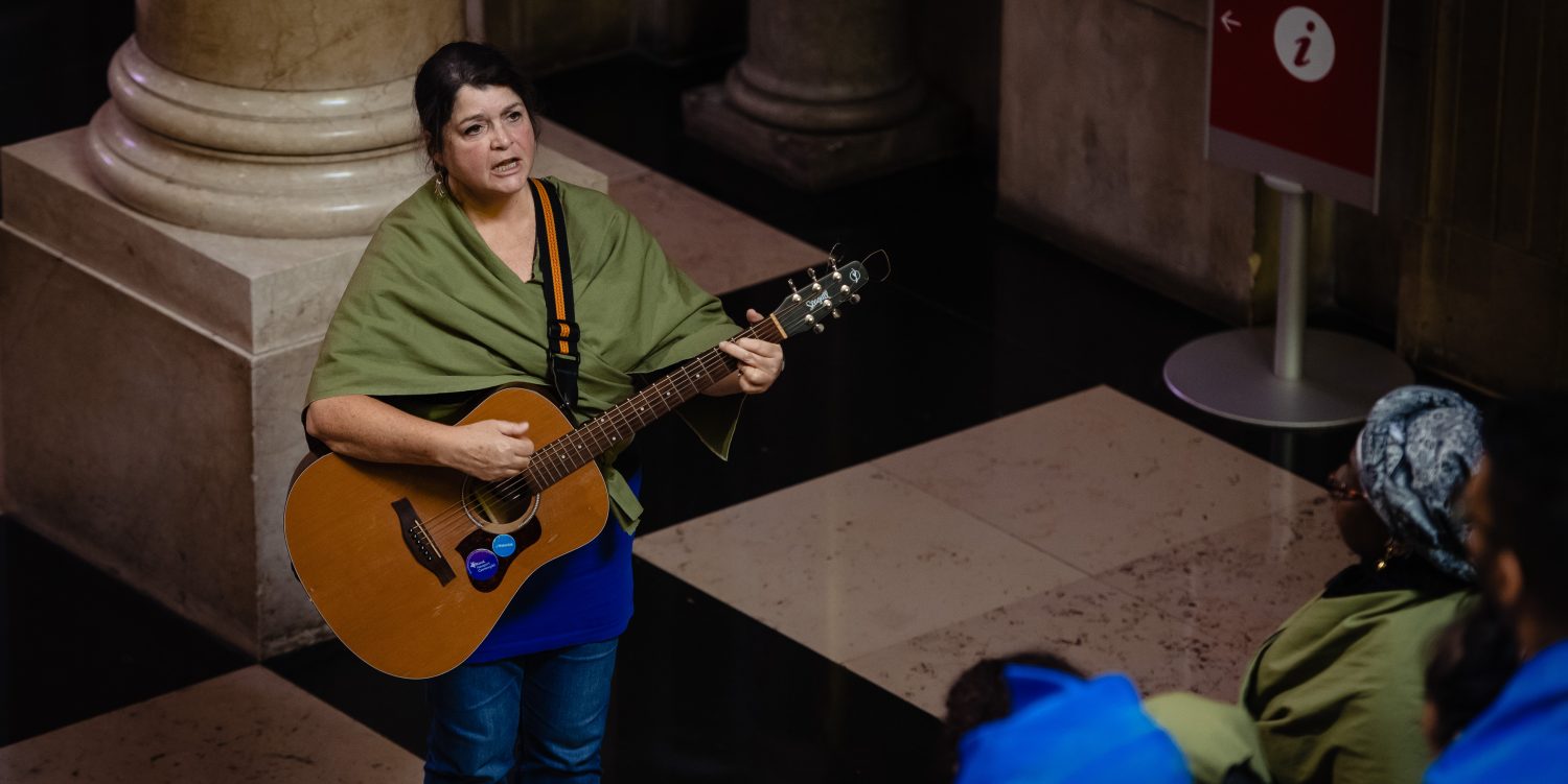 A woman is playing a guitar and singing a song, facing a choir of singers.