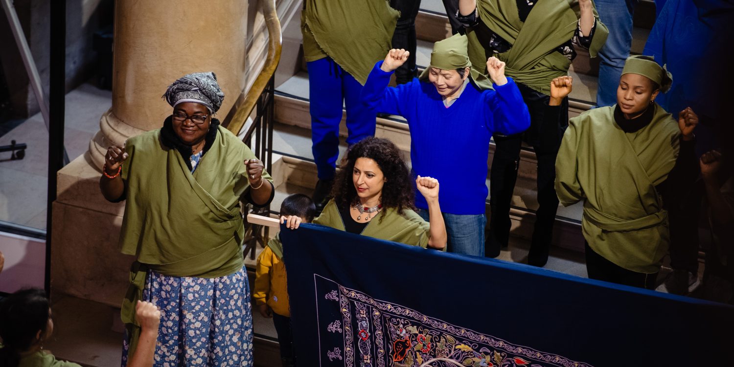 People who form a choir wearing khaki and blue clothes are stood smiling, holding up both hands in a fist as a sign of solidarity.