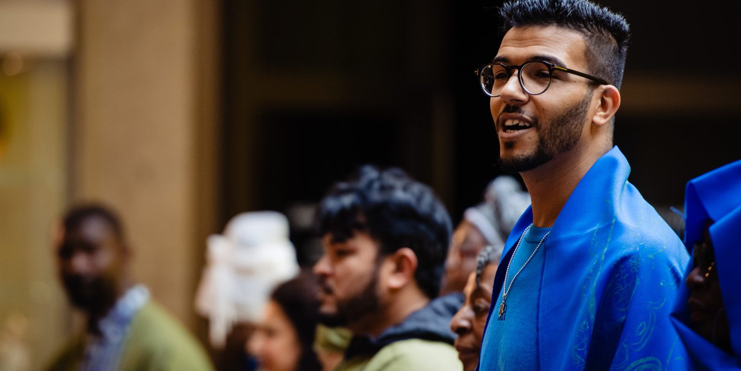 In focus is a man with short dark hair, wearing glasses and blue clothes. He is mid-song. Behind him and out of focus are other members of a choir.