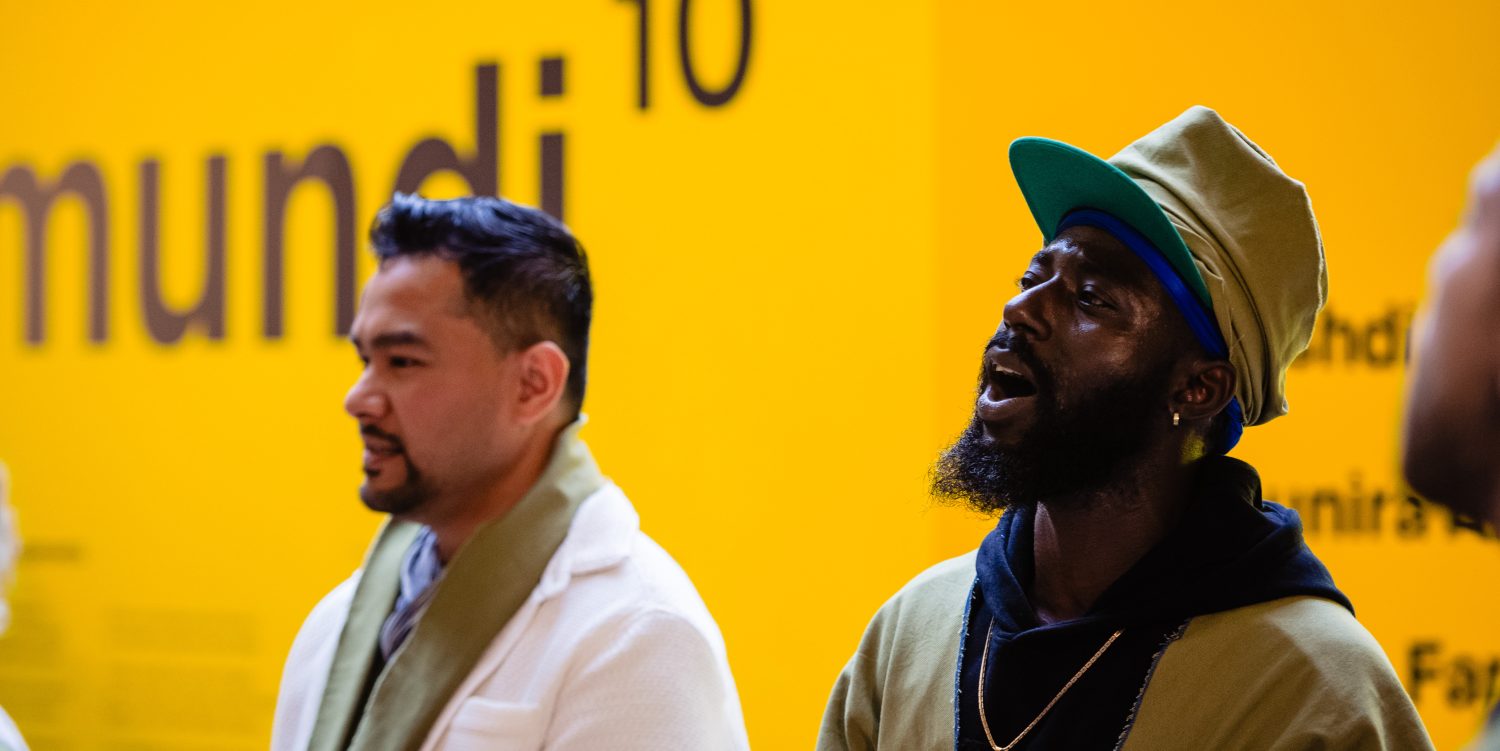 A man in focus, wearing a cap, is mid-song. Next to him and slightly out of focus is another man in a white blazer. They are stood in front of a yellow wall that reads 'Artes Mundi 10'.