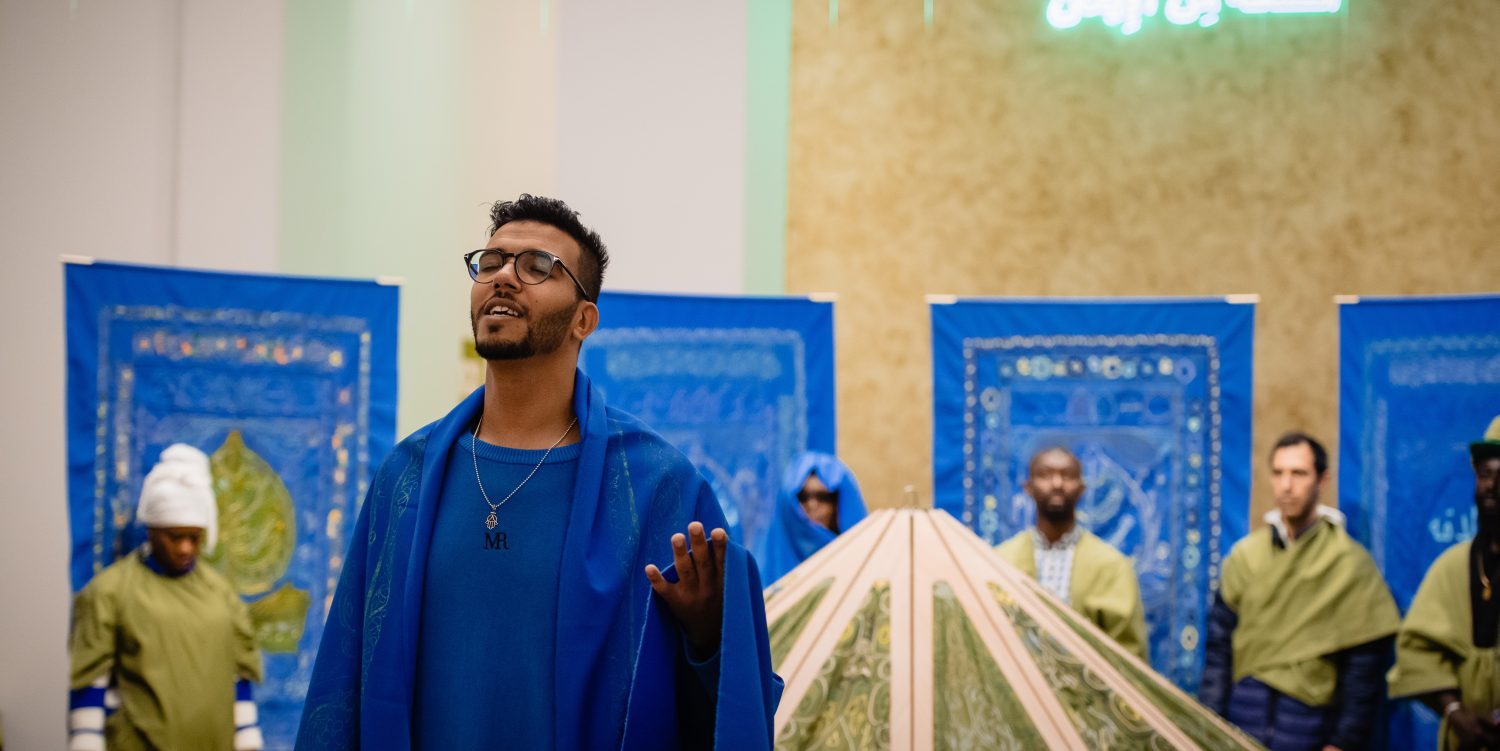 A man in blue robes is singing and using his hand to gesture. Behind him is a row of people, standing in front of blue tapestries, and behind an embroidered tent.