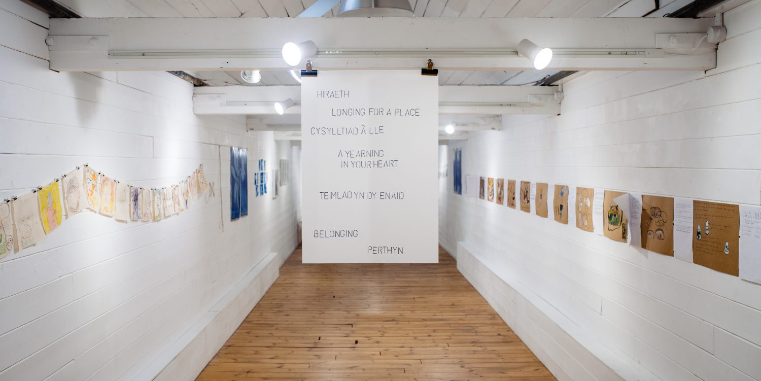 A gallery with prints and artwork are displayed on the wall. Hung in the centre of a room is a white sheet which reads: HIRAETH LONGING FOR A PLACE CYSYLLTIAD A LLE A YEARNING IN YOUR HEART TEIMLAD YN DY ENAID BELONGING PERTHYN