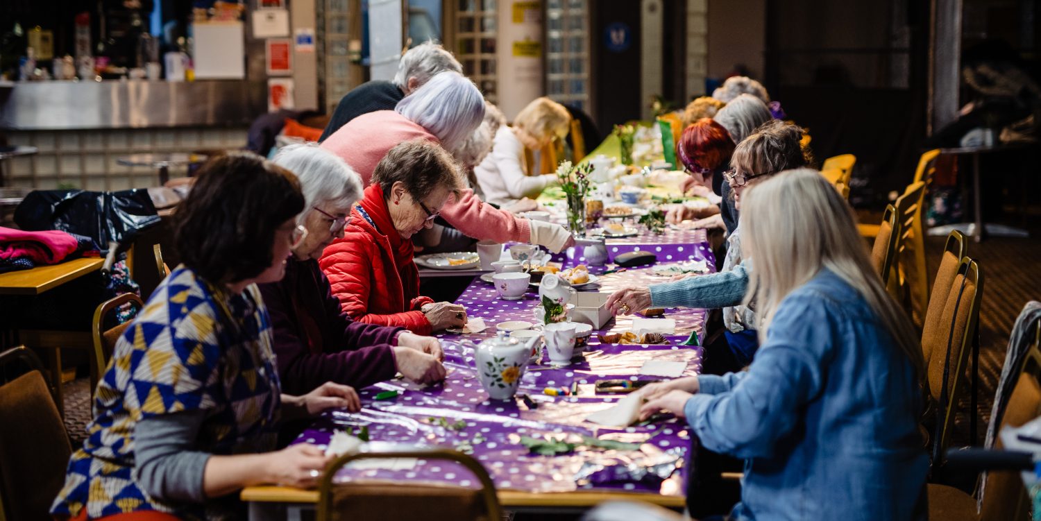 Elderly ladies are sat down at a long table covered in a purple tablecloth with white polka dots. They are using leaves and natural items to create prints.