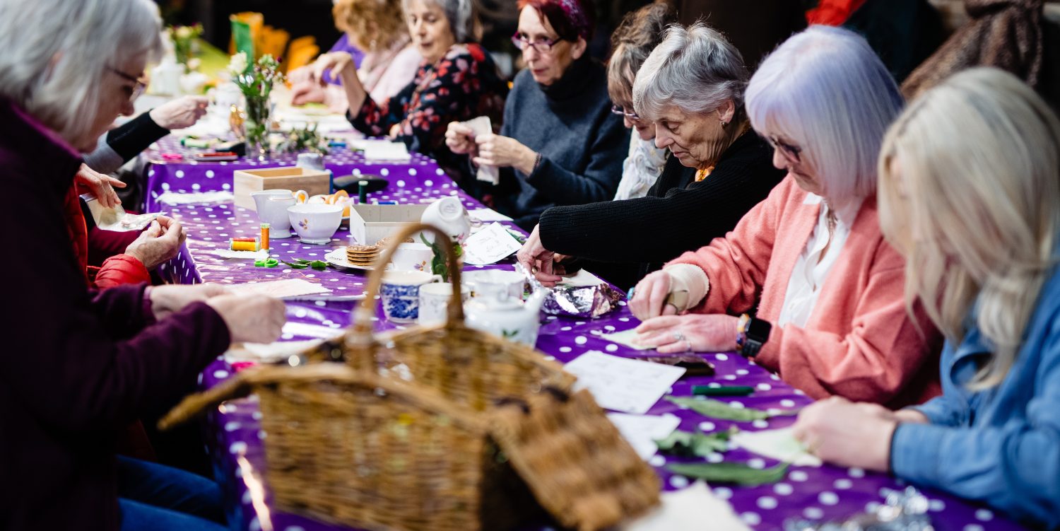 Elderly ladies are sat down at a long table covered in a purple tablecloth with white polka dots. They are using leaves and natural items to create prints.