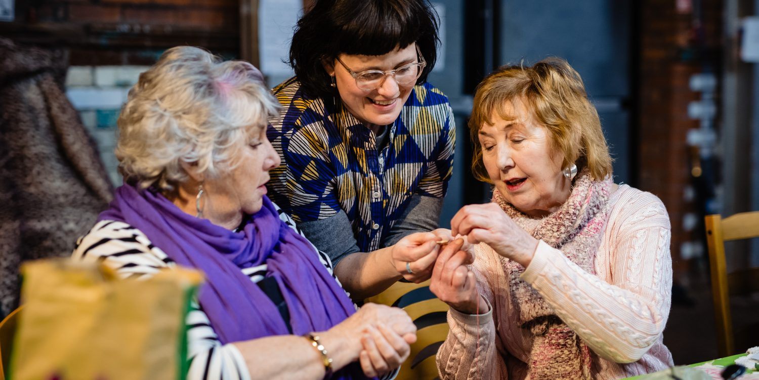 Two elderly ladies are sat down at a table, talking to a female workshop leader and getting help with their embroidery.