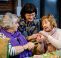 Two elderly ladies are sat down at a table, talking to a female workshop leader and getting help with their embroidery.