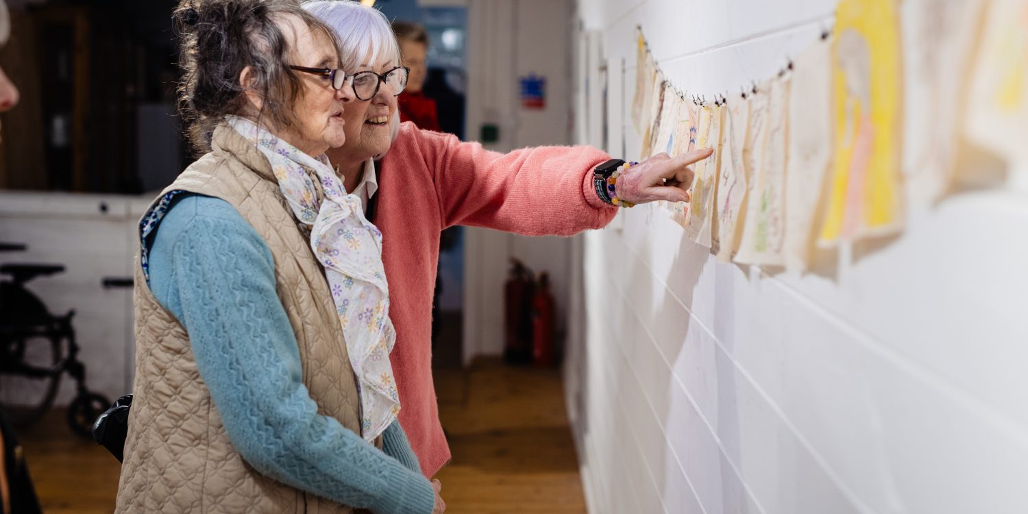 Two elderly ladies are looking and pointing at embroidered pieces of fabric hung up on a gallery wall.