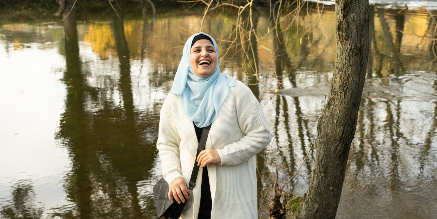 A woman wearing a long cream cardigan and a pale blue hijab is laughing and standing next to a river.