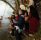 Parents with young children are stood at the side of a river pointing and smiling. The father is holding a bunch of willow as they are on a willow raft workshop,