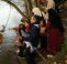 Parents with young children are stood at the side of a river pointing and smiling. The father is holding a bunch of willow as they are on a willow raft workshop,