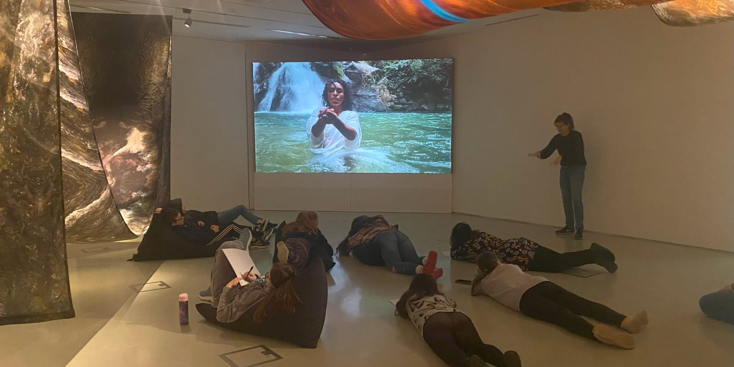 People are lying on the floor or on beanbags in a gallery room. There is a film on a screen in the corner of the gallery and draped fabric can be seen in other areas of the gallery.