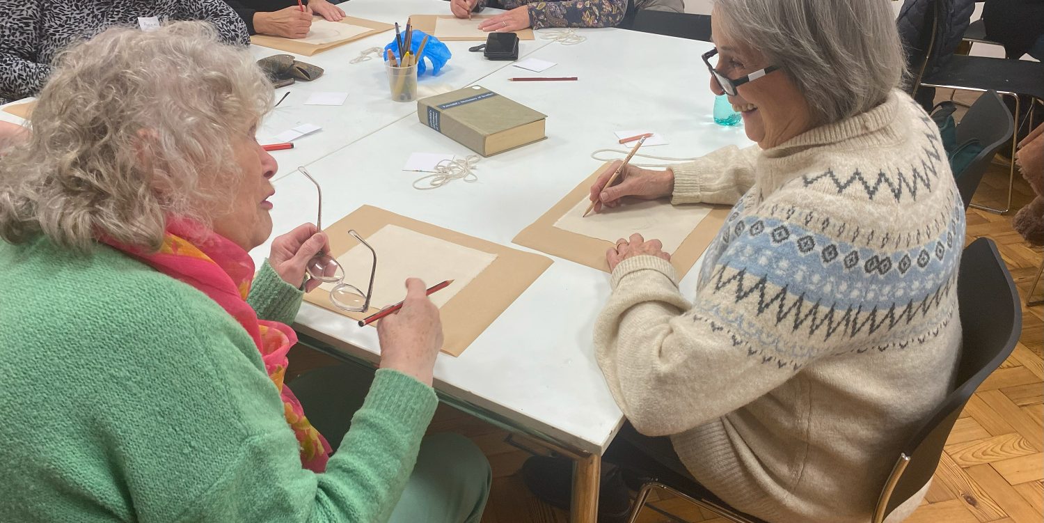 Two elderly women, wearing winter jumpers are drawing with pencils at a table. They are talking and laughing together.