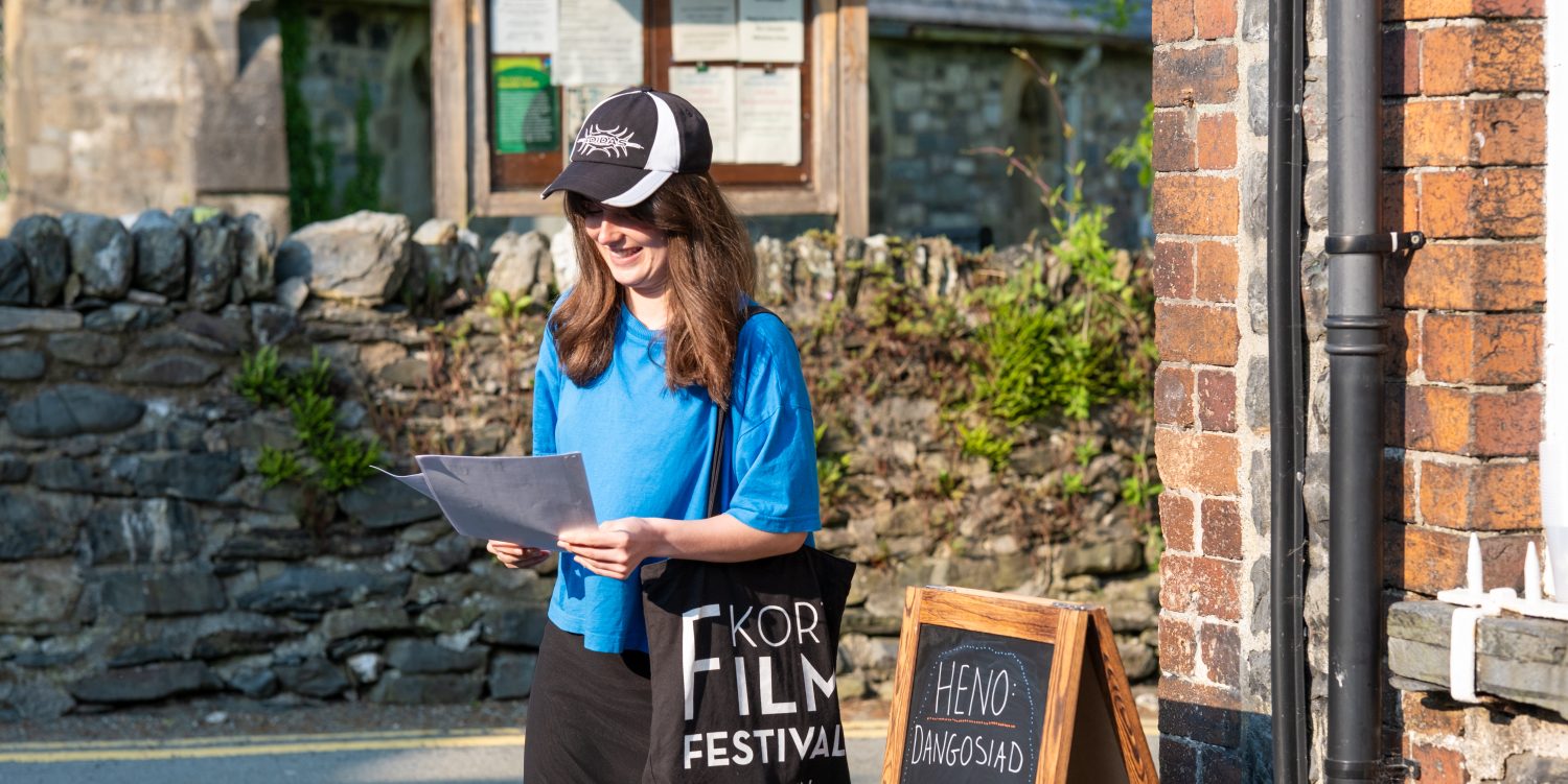 A woman wearing a black and white cap, blue t-shirt, and black skirt is holding A4 sheets of paper outside of the gallery, Oriel Machno.