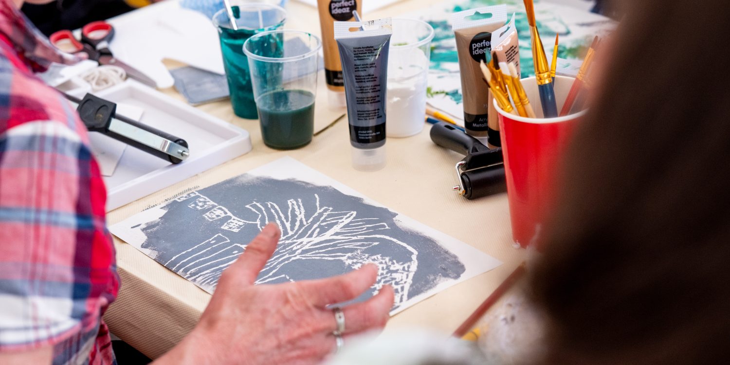 A person is sat down looking at the monoprint they have just created. On the table is paint, rollers, paint brushes and pots of water.