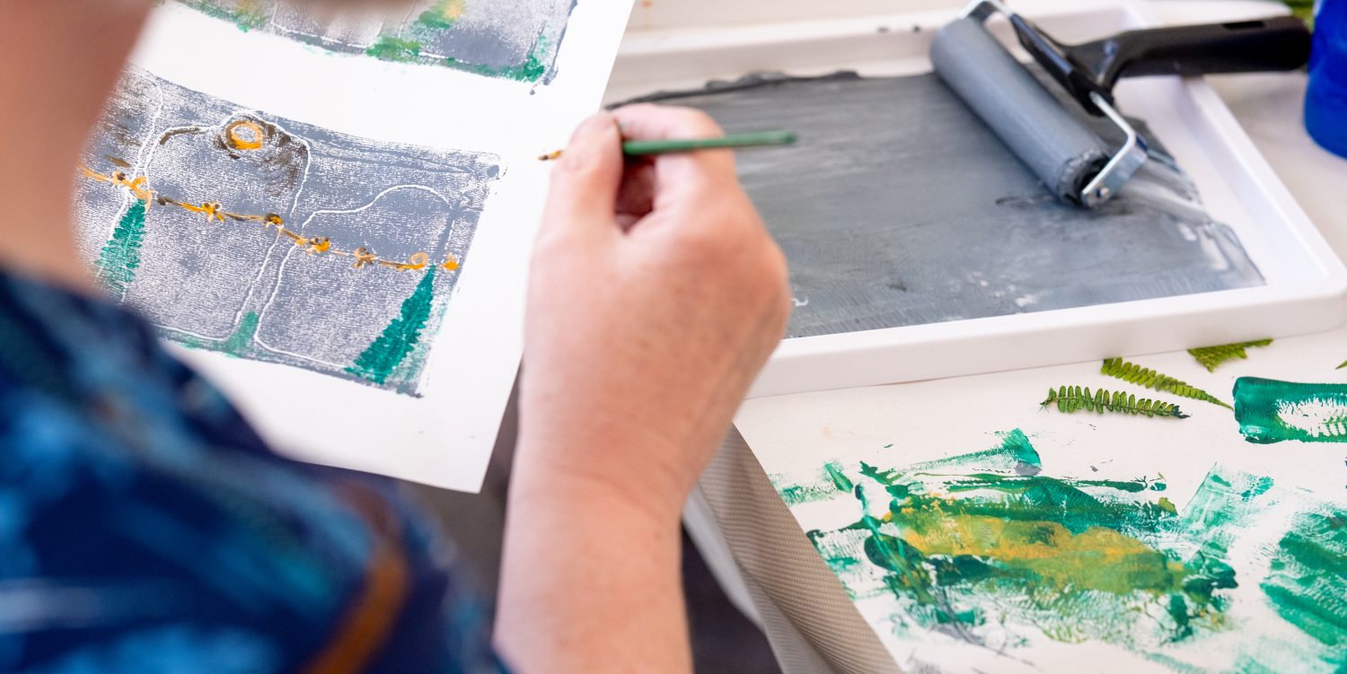 In the foreground, a person is out of focus. The print they are holding up is in focus. In their other hand they are holding a small paintbrush. In the background you can see paint on a tray and a roller.