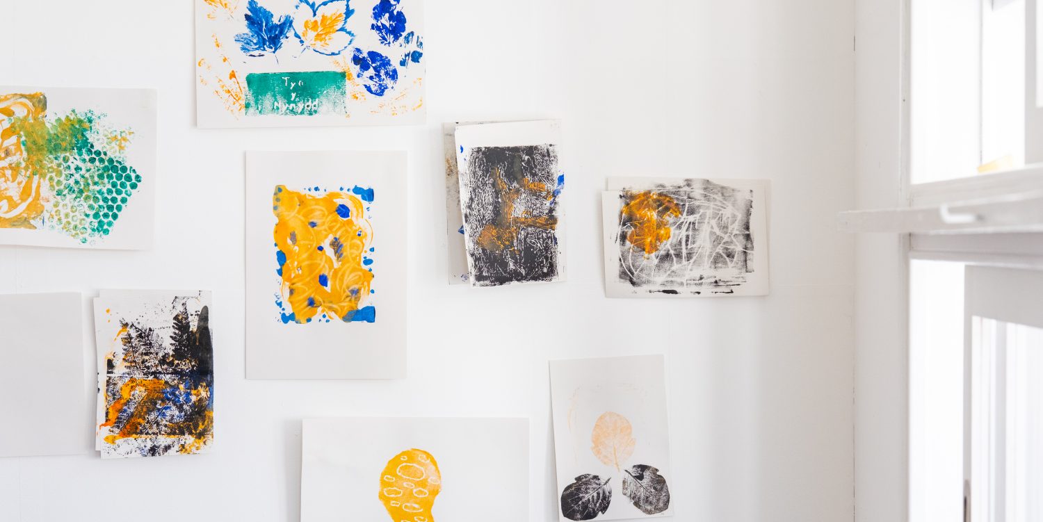 Individual monoprints are stuck up on a plain white wall.