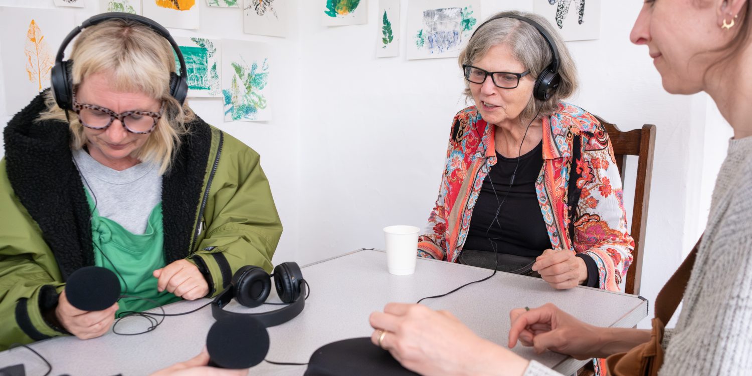 Three women are sat around a table, looking at and discussing sound recording equipment including microphones and headphones.