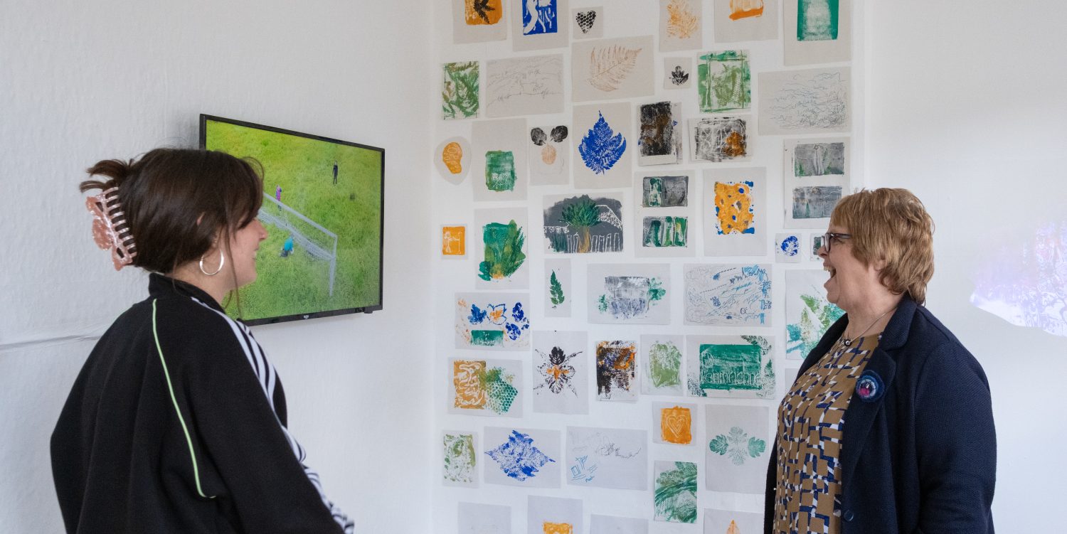 Two people in a gallery room are stood in front of a wall of monoprints chatting and laughing together.