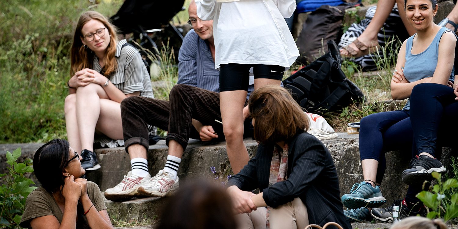 As part of a spoken word collaborative performance, a woman stands on steps holding embroidered fabric and looking towards another person facing away from the camera. Around them, others sit on various steps, watching the two people standing.