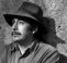 A man with a moustache wearing a hat and collared shirt sits against a textured wall. He has a slight smile and is looking away from the camera. It is a black and white image.