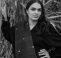 A woman with long hair poses for a portrait photo in front of long-leaved plants. She has one hand on her hip and the other holds on to the plant. She has a decorative scarf draped over her shoulder. It is a black and white image.