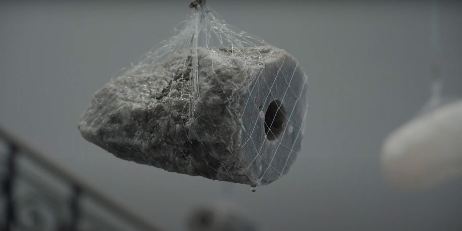 A roughly cut piece of salt is suspended in a net high above the ground, with a water droplet poised to fall from it.