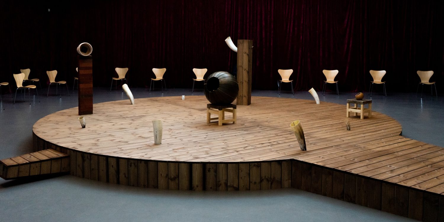 A raised circular wooden platform features sculptures that are part of an experimental opera. The sculptures play opera music, and chairs are arranged around the platform for visitors to experience the installation.