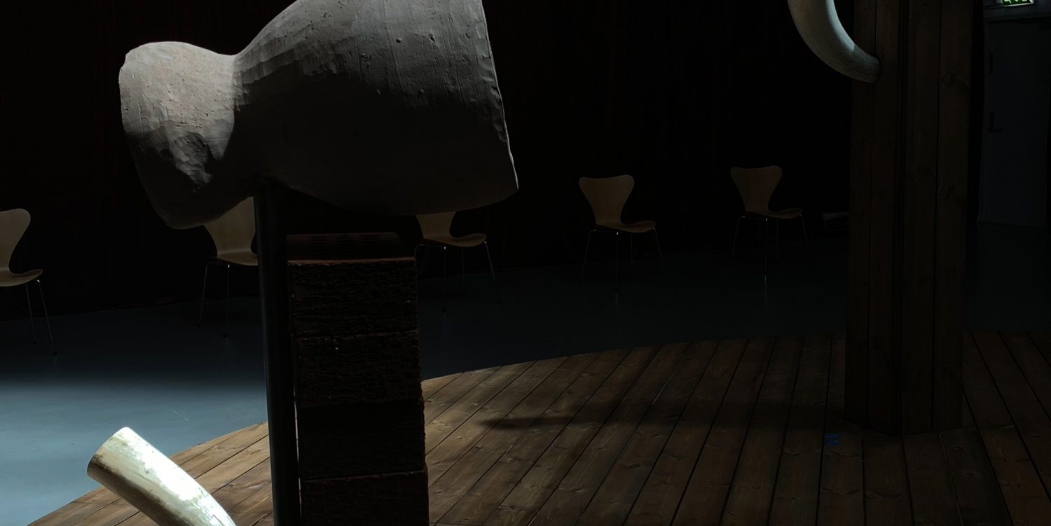 On a raised wooden platform, three sculptures are displayed as part of an experimental opera installation. The sculptures emit opera music, and surrounding the platform are chairs for viewers to sit and experience the piece.