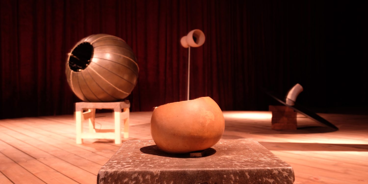 On a raised wooden platform, four sculptures are arranged. In the foreground is a circular sculpture on a stool, while three additional sculptures are positioned in the background. This setup is part of an experimental opera where the sculptures produce opera music.